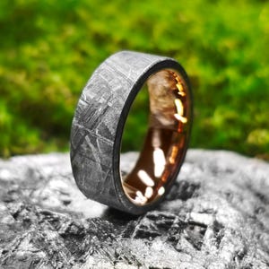 May include: A close-up of a ring with a patterned, dark gray exterior and a gold-colored interior. The ring is set against a blurred green and gray background, highlighting the intricate design and metallic textures.