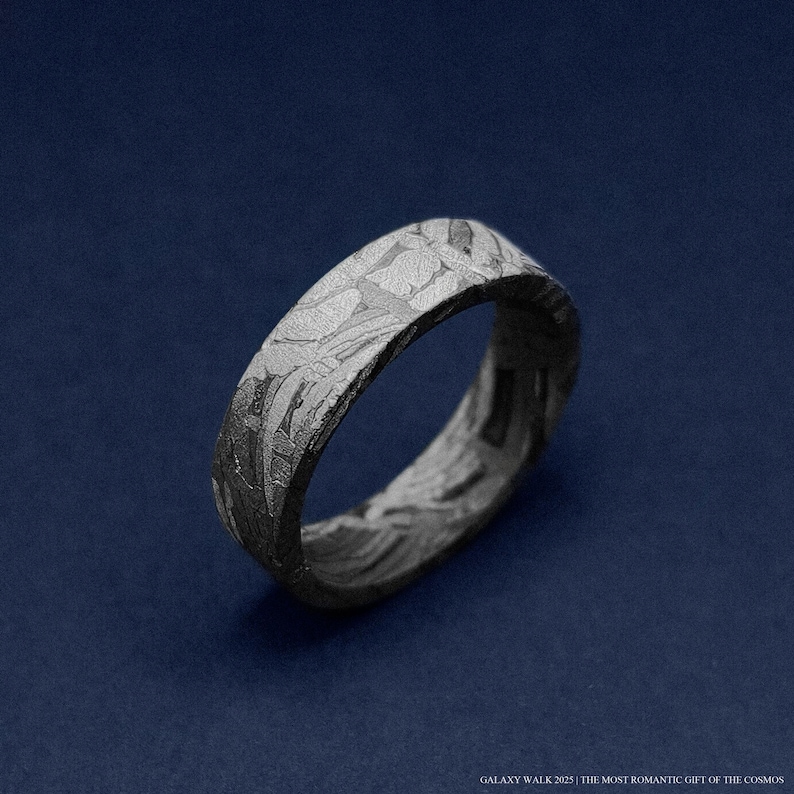 K&ouml;nnte beinhalten: Nahaufnahme eines strukturierten, dunkelgrauen Rings mit einem einzigartigen Muster. Der Ring ist vor einem dunkelblauen Hintergrund platziert. Der Text unten lautet "GALAXY WALK 2025 | THE MOST ROMANTIC GIFT OF THE COSMOS."