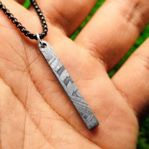 Puede incluir: Un collar con colgante plateado con un colgante de barra rectangular. El colgante tiene un diseño único y está suspendido de una cadena negra. El collar se muestra sobre un fondo color piel.