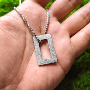 Puede incluir: Un collar de cadena de plata con un colgante rectangular hecho de meteorito. El colgante tiene un recorte rectangular en el centro.