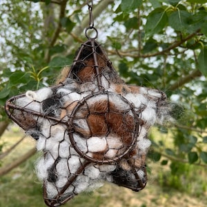 May include: A star-shaped hanging planter with a wire frame and filled with a mix of white, brown, and black wool. The planter is suspended by a metal chain, perfect for displaying small plants or flowers. The star is approximately 8 inches wide.