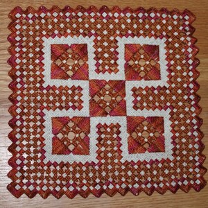Op de afbeelding: Vierkant, handgestikt textiel met een geometrisch ontwerp. Het stuk heeft een centraal kruisachtig patroon in bruin- en oranje tinten, omgeven door een witte achtergrond en een geschulpte rand. Afmetingen: ongeveer 20 cm bij 20 cm.