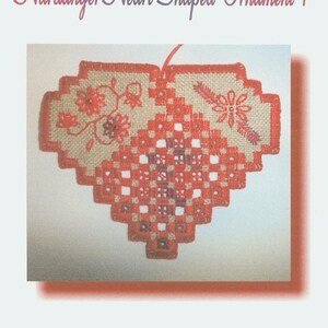 Op de afbeelding: Een hartvormige Hardanger-borduurwerk ornament met rode en witte steken op een beige stof. De ornament heeft een bloemmotief en de tekst "Hardanger Heart-Shaped Ornament 1". Ontworpen door April Morgan-Salo.