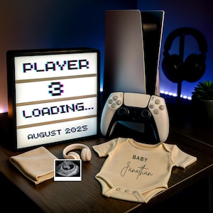 Op de afbeelding: Een baby aankondiging met een gaming thema. Een lichtbak toont "PLAYER 3 LOADING... AUGUST 2025." Een witte game controller, een spelconsole en een baby rompertje met de naam "Jonathan" zijn ook afgebeeld.