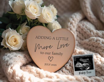 May include: A wooden heart-shaped sign with the text "Adding a little more love to our family" and the date "July 2025". A black and white ultrasound image is in the background with the text "Baby Rose".