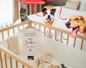 May include: A white baby onesie with the text "LITTLE SECRET IS ON THE WAY BABY Janothan JULY 2027" hangs in a wooden crib. The crib contains a blanket, booties, a hat, and a wooden leaf. An ultrasound image is also present.
