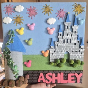May include: A square wall art piece featuring a light blue sky with pink and gold firework cutouts, white clouds, and colourful mouse-shaped icons. It displays a grey stone castle, a blue-roofed tower with green vines, and the name ASHLEY.