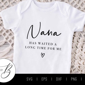 May include: White baby onesie with black text that reads "Nana has waited a long time for me" with a small heart symbol below the text.