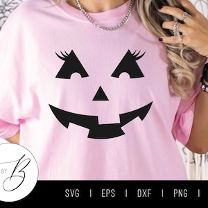 May include: A pink t-shirt featuring a black jack-o'-lantern face graphic. The graphic depicts a smiling jack-o'-lantern with eyelashes, a triangular nose, and a wide grin.