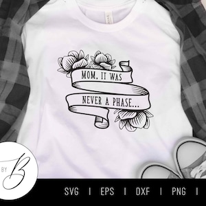 Könnte beinhalten: Weißes T-Shirt mit einem schwarzen floralen Banner-Design mit dem Text "MOM, IT WAS NEVER A PHASE...". Das Shirt wird mit einem schwarz-grauen karierten Hemd und grauen Leinenschuhen präsentiert.