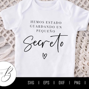 May include: White baby onesie with the Spanish text "HEMOS ESTADO GUARDANDO UN PEQUEÑO Secreto" in black script. A small heart is below the word "Secreto". The onesie is on a white and beige textured surface.