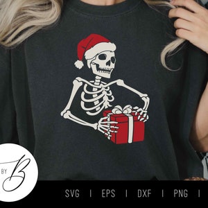 Puede incluir: Una camiseta negra con un esqueleto blanco que lleva un gorro de Papá Noel rojo y sostiene una caja de regalo roja con un lazo blanco.