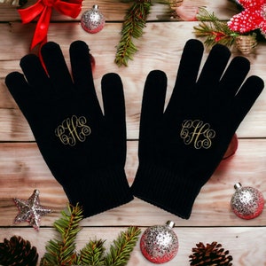 May include: A pair of black knit gloves with gold embroidered initials "GHS" on each glove. The gloves are photographed on a wooden surface with Christmas decorations.
