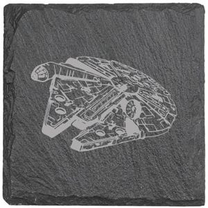 May include: A square, dark gray slate coaster featuring a detailed, light gray etched image of the Millennium Falcon starship. The coaster has a rough, textured surface and is designed for use with beverages.