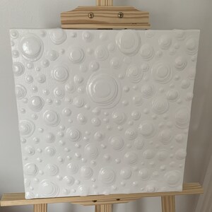 May include: White canvas painting with a textured surface of raised circles and rings. The painting is on an easel.