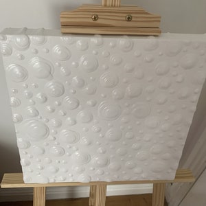 May include: White canvas painting with a textured surface of raised circles. The painting is on an easel.