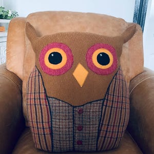 May include: A brown owl-shaped pillow with a plaid vest and button details. The owl has large yellow eyes with black pupils and a yellow beak.