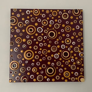 May include: A square canvas painting with a maroon background and a pattern of gold and white circles. Some of the circles have a concentric ring pattern.