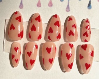 May include: Set of ten nude-colored press-on nails with a red heart pattern. The nails are displayed on a white card with the text "Dreams By Maya".