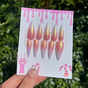May include: Set of ten pink and white ombre stiletto false nails with a pearlescent finish. The nails are displayed on a white card with pink dripping paint and the text "Claws By Maya".