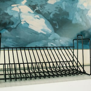 May include: A black metal wire rack featuring a series of evenly spaced vertical dividers designed for organizing items. The structure has a rectangular base with a slanted frame and two taller vertical supports on each side for stability.
