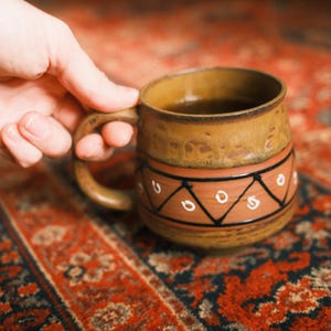 Może przedstawiać: Brązowy ceramiczny kubek z uchwytem, wypełniony ciemnym płynem. Kubek ma ozdobny pas z geometrycznymi wzorami w kolorze czarnym i białym. Kubek jest trzymany przez dłoń, spoczywający na czerwonym, wzorzystym dywanie.