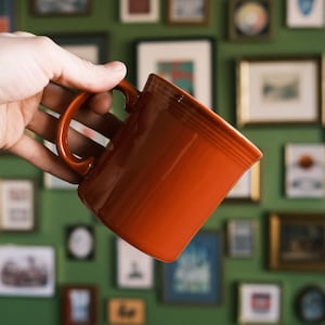 Peut inclure: Une tasse en céramique orange vif avec une anse incurvée est tenue. La tasse a une finition brillante et un design classique. L'arrière-plan présente une collection d'images et d'œuvres d'art encadrées.