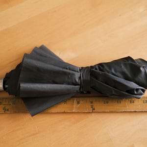 Puede incluir: Un paraguas negro plegado con un mango plateado y una correa de mu&ntilde;eca negra. El paraguas est&aacute; sobre una regla de madera, que mide hasta 40 cm. El paraguas est&aacute; asegurado con una correa negra.