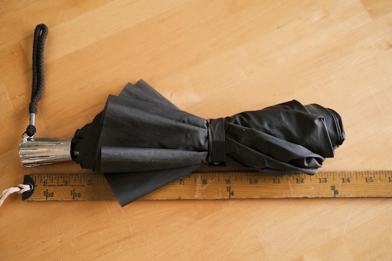 Puede incluir: Un paraguas negro plegado con un mango plateado y una correa de mu&ntilde;eca negra. El paraguas est&aacute; sobre una regla de madera, que mide hasta 40 cm. El paraguas est&aacute; asegurado con una correa negra.