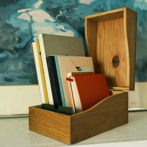 May include: A wooden box with a hinged lid, containing several books and notebooks. The books are various colors, including orange, gray, and black. The box is made of light-colored wood and sits on a white surface.