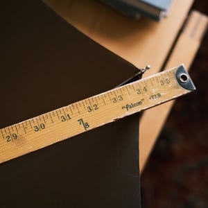 Puede incluir: Una regla de madera, marcada en pulgadas, se encuentra en diagonal sobre una superficie marr&oacute;n oscuro. La regla, etiquetada como "Falcon" y "72R", muestra medidas de 68.58 cm a 88.9 cm. La imagen sugiere un contexto artesanal o de medici&oacute;n.