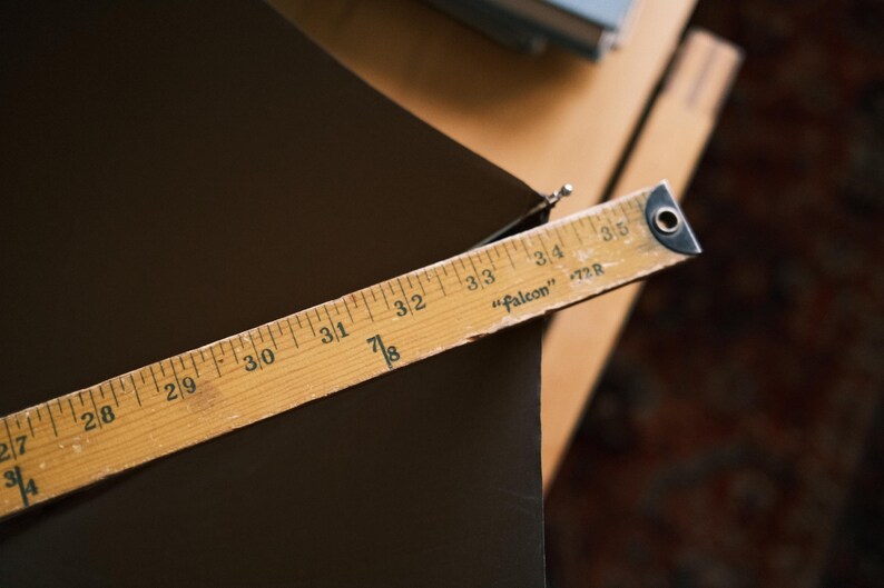 Puede incluir: Una regla de madera, marcada en pulgadas, se encuentra en diagonal sobre una superficie marr&oacute;n oscuro. La regla, etiquetada como "Falcon" y "72R", muestra medidas de 68.58 cm a 88.9 cm. La imagen sugiere un contexto artesanal o de medici&oacute;n.