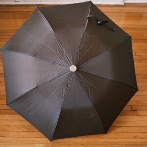 Puede incluir: Un paraguas negro, completamente abierto, con un bot&oacute;n plateado en el centro. La tela del paraguas es negra y tiene un asa en la parte superior. El paraguas est&aacute; sobre un suelo de madera.