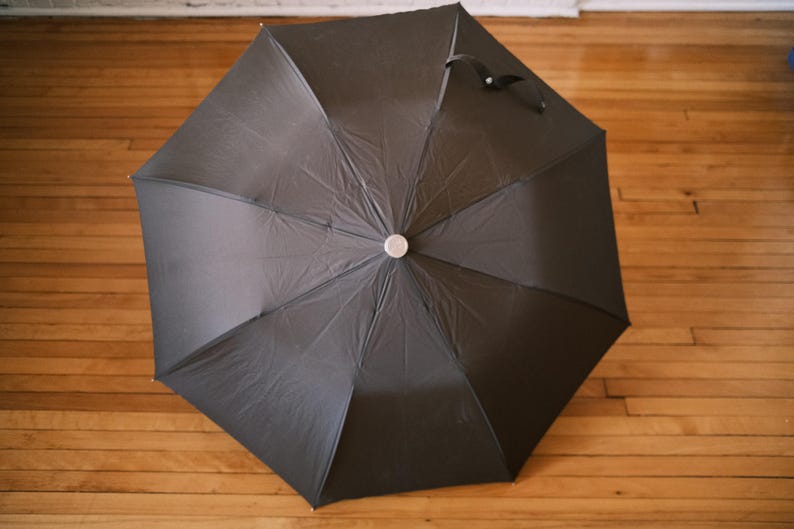 Puede incluir: Un paraguas negro, completamente abierto, con un bot&oacute;n plateado en el centro. La tela del paraguas es negra y tiene un asa en la parte superior. El paraguas est&aacute; sobre un suelo de madera.