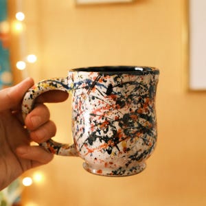Puede incluir: Una taza de cerámica con base blanca que presenta un patrón de salpicaduras de pintura multicolor en negro, naranja y azul. La taza tiene un cuerpo redondeado, un asa resistente y un acabado liso y brillante. El interior de la taza es oscuro.