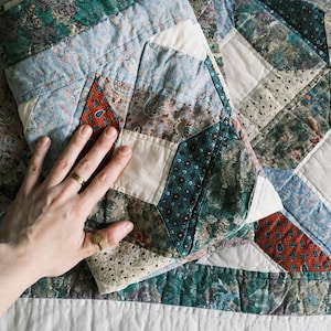 Puede incluir: Un edredón hecho a mano con un diseño de patchwork. El edredón presenta varias telas en tonos de azul, verde, rojo y blanco. El edredón cosido a mano tiene un patrón geométrico y un borde blanco. El edredón está doblado.
