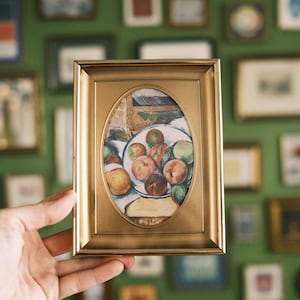 May include: A rectangular gold-toned picture frame with an oval-shaped center opening. The frame contains a print of a still life painting featuring a white bowl filled with various round fruits, including peaches and green apples, on a table.