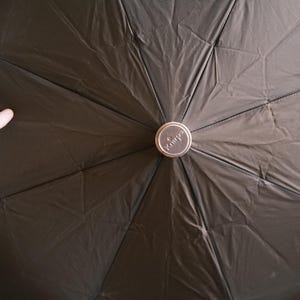 Puede incluir: Un paraguas negro con un bot&oacute;n central plateado. La tela del paraguas tiene un aspecto texturizado. La palabra "Knirps" es visible en el bot&oacute;n. El paraguas est&aacute; abierto.