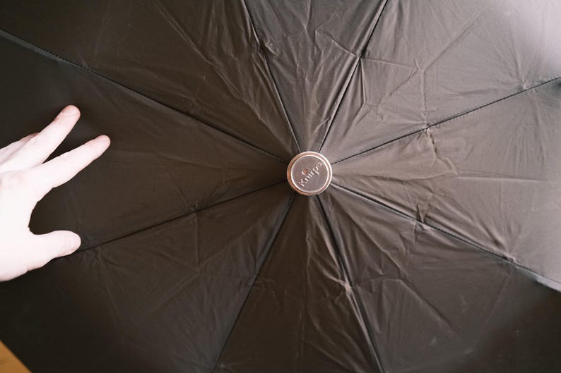 Puede incluir: Un paraguas negro con un bot&oacute;n central plateado. La tela del paraguas tiene un aspecto texturizado. La palabra "Knirps" es visible en el bot&oacute;n. El paraguas est&aacute; abierto.