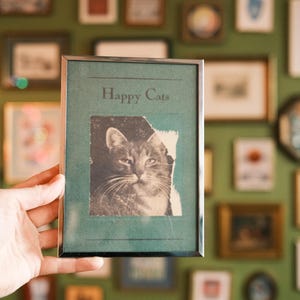 May include: A silver-framed print featuring a monochrome image of a cat and the words "Happy Cats". The print is held against a wall adorned with various framed pictures.