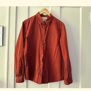 Puede incluir: Una camisa de manga larga de color óxido con un solo bolsillo en el pecho. La camisa tiene un logotipo de Columbia en el lado izquierdo del cuello.