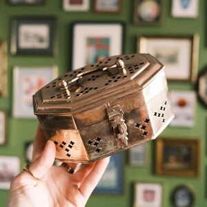 May include: A vintage brass box with an octagonal shape, a handle, and a latch. The box has decorative cut-out patterns and is held against a backdrop of framed pictures on a green wall.