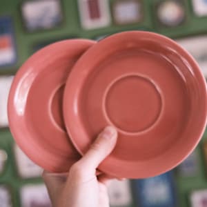May include: Two round, solid coral-colored ceramic saucers with a smooth, glossy finish. Each saucer features a slightly raised outer rim and a central circular indentation designed to hold a cup securely in place, showcasing a uniform color.