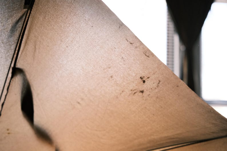 Puede incluir: Primer plano de una tela beige con manchas oscuras y un contorno negro. La tela est&aacute; tensa, creando una forma triangular. La textura parece ligeramente &aacute;spera, con imperfecciones visibles.