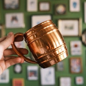 May include: A copper Moscow mule mug with a hammered finish and a rounded handle. The mug is being held in a hand against a background of framed pictures.