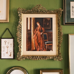 May include: A collection of framed art pieces hangs on a green wall. The central piece is a framed painting of a woman in a rust-colored dress. Other frames include abstract art and a framed print of keys.