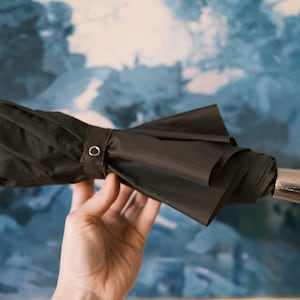 Puede incluir: Un paraguas negro compacto con un mango plateado y una correa de mu&ntilde;eca negra. El paraguas est&aacute; plegado y asegurado con una banda de tela a juego y un broche met&aacute;lico. El fondo es azul y blanco borroso.