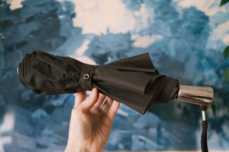 Puede incluir: Un paraguas negro compacto con un mango plateado y una correa de mu&ntilde;eca negra. El paraguas est&aacute; plegado y asegurado con una banda de tela a juego y un broche met&aacute;lico. El fondo es azul y blanco borroso.