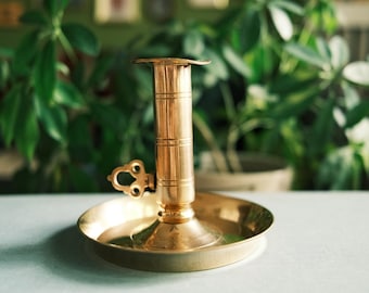 May include: A polished brass chamberstick candle holder featuring a wide circular drip tray base, a central cylindrical stem with horizontal engraved lines, and a small decorative handle loop attached to the side for easy carrying.