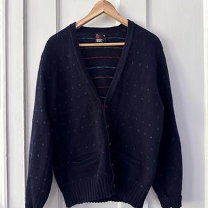 Könnte beinhalten: Ein marineblauer Cardigan mit einem kleinen weißen Tupfenmuster. Der Cardigan hat einen V-Ausschnitt, lange Ärmel und zwei Taschen auf der Vorderseite. Der Cardigan hängt an einem Holzbügel. Das Etikett auf dem Cardigan lautet "Robert Bruce".